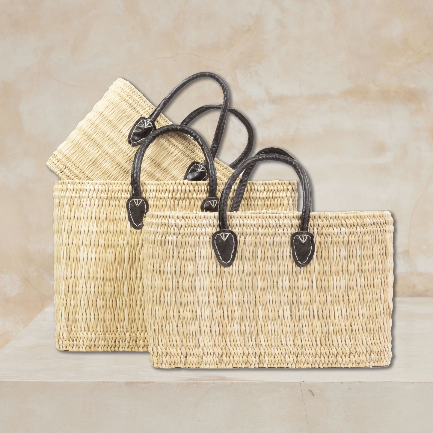 three woven straw tote bags with dark brown leather handles, displayed on a light beige stone pedestal against a textured beige wall.